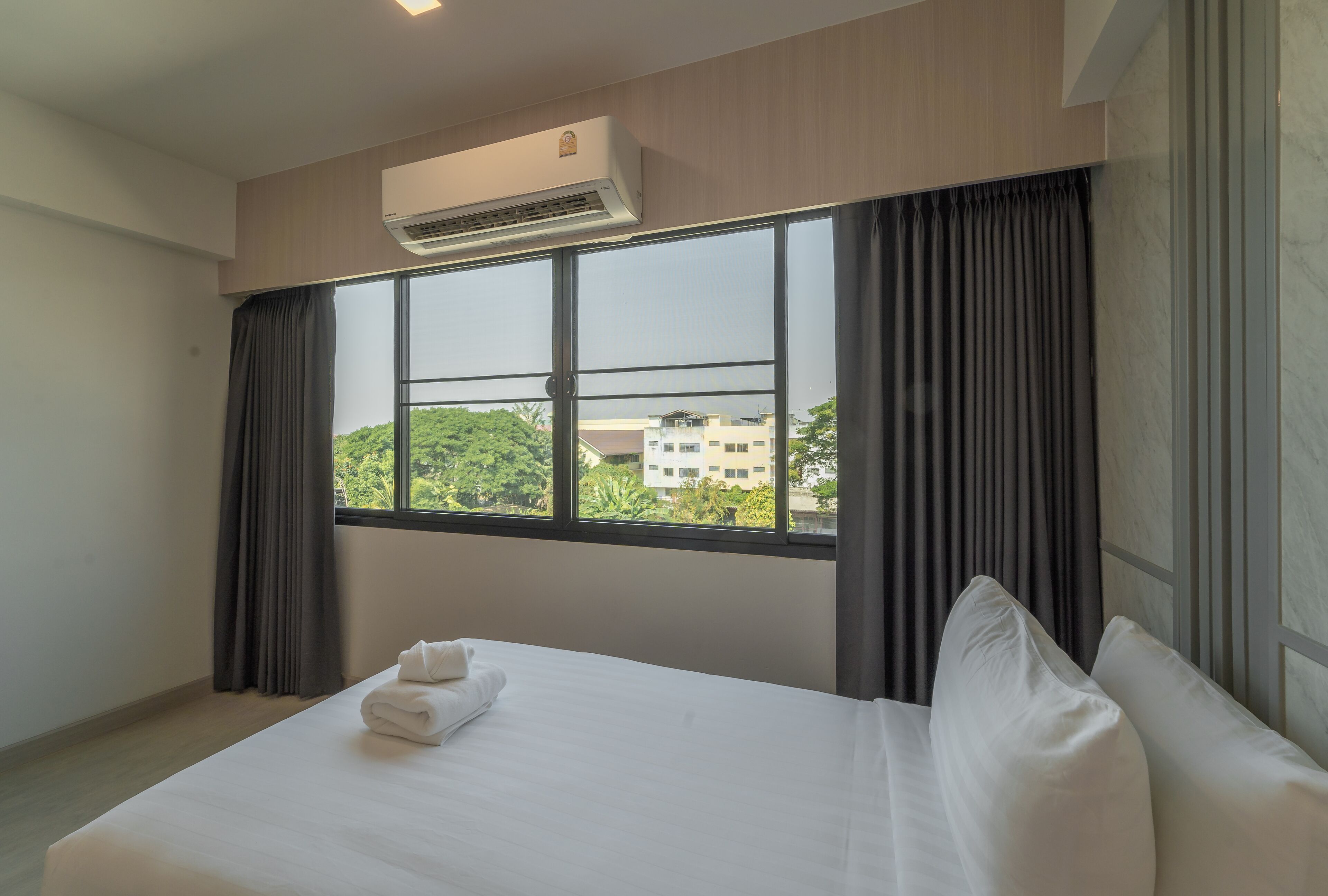 A hotel room with a large bed, a window with curtains, an air conditioning unit, and a view of buildings and greenery outside.