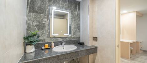 Une salle de bain moderne avec un lavabo en marbre, un miroir doté d’un éclairage intégré, une plante en pot et une poubelle.