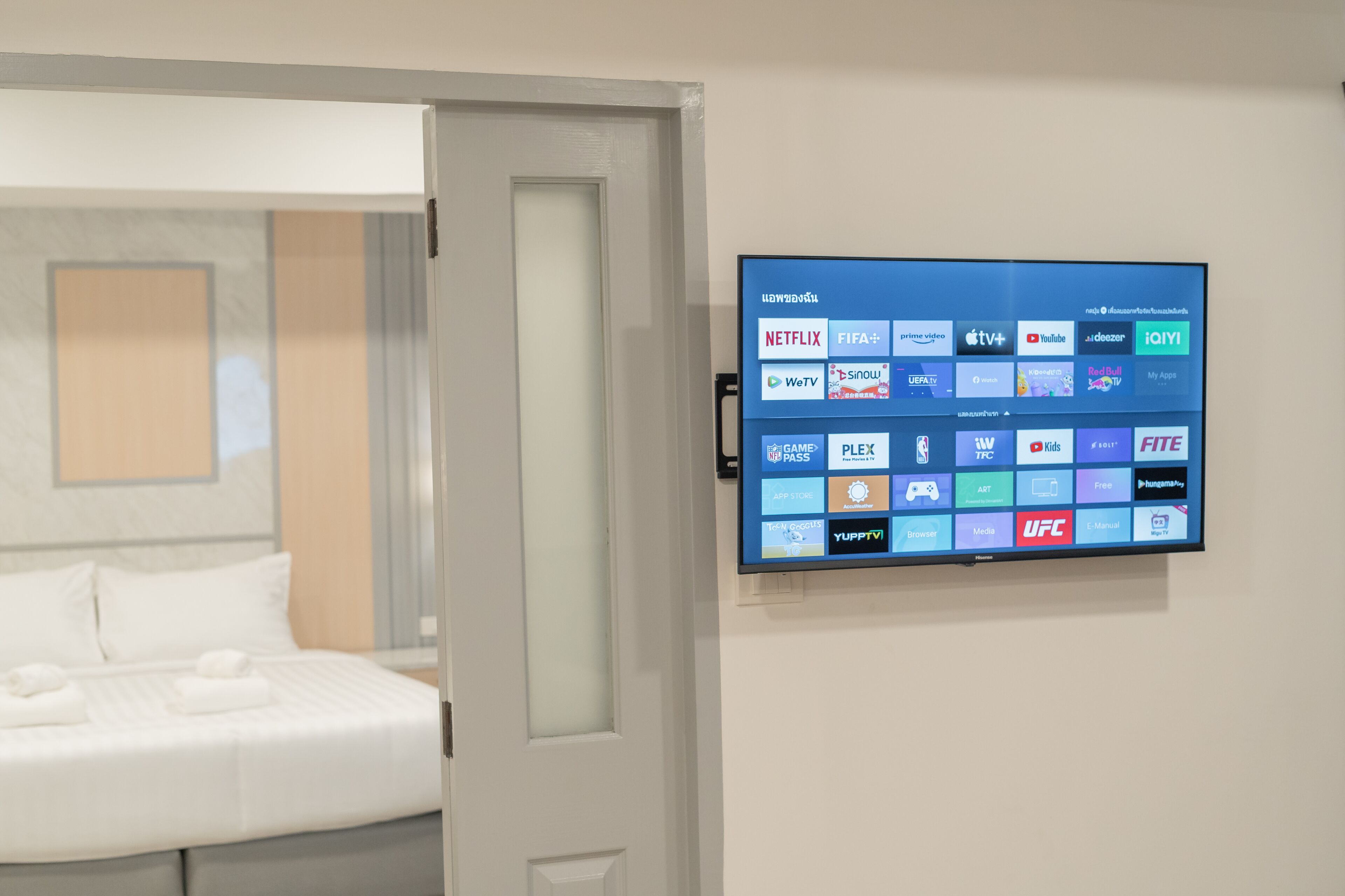 A hotel room with a bed, a flat-screen TV mounted on the wall displaying various streaming services, and a door leading to another room.