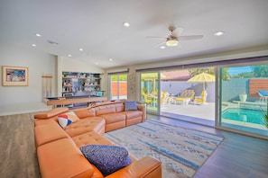 A modern living room with a sectional sofa, a kitchen area, and a view of a backyard with a pool.