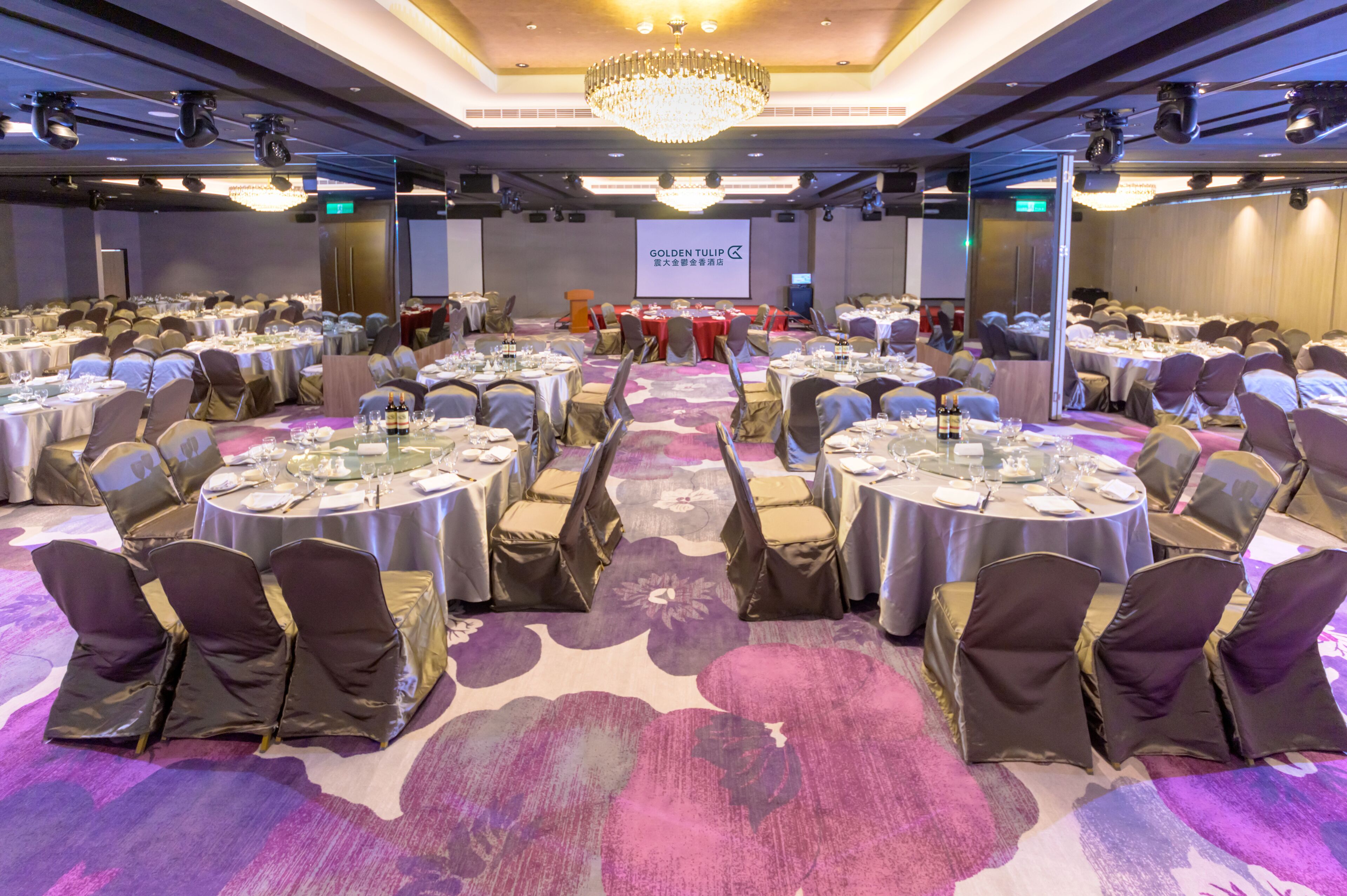 Salão de banquetes com mesas redondas, cadeiras e palco com uma tela exibindo "GOLDEN TULIP".