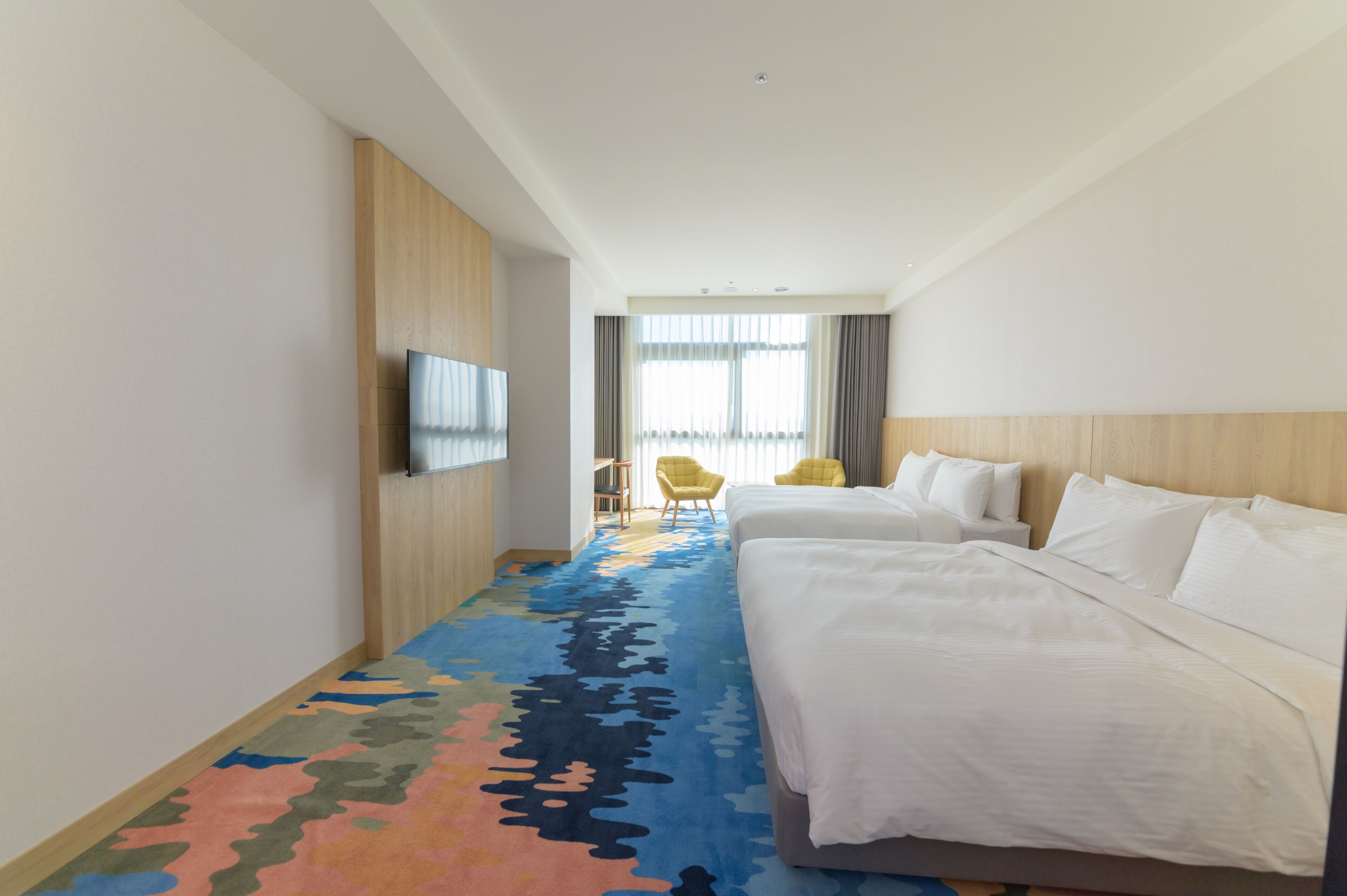 A hotel room with a large bed, a TV mounted on the wall, a colorful carpet, and a window with curtains.