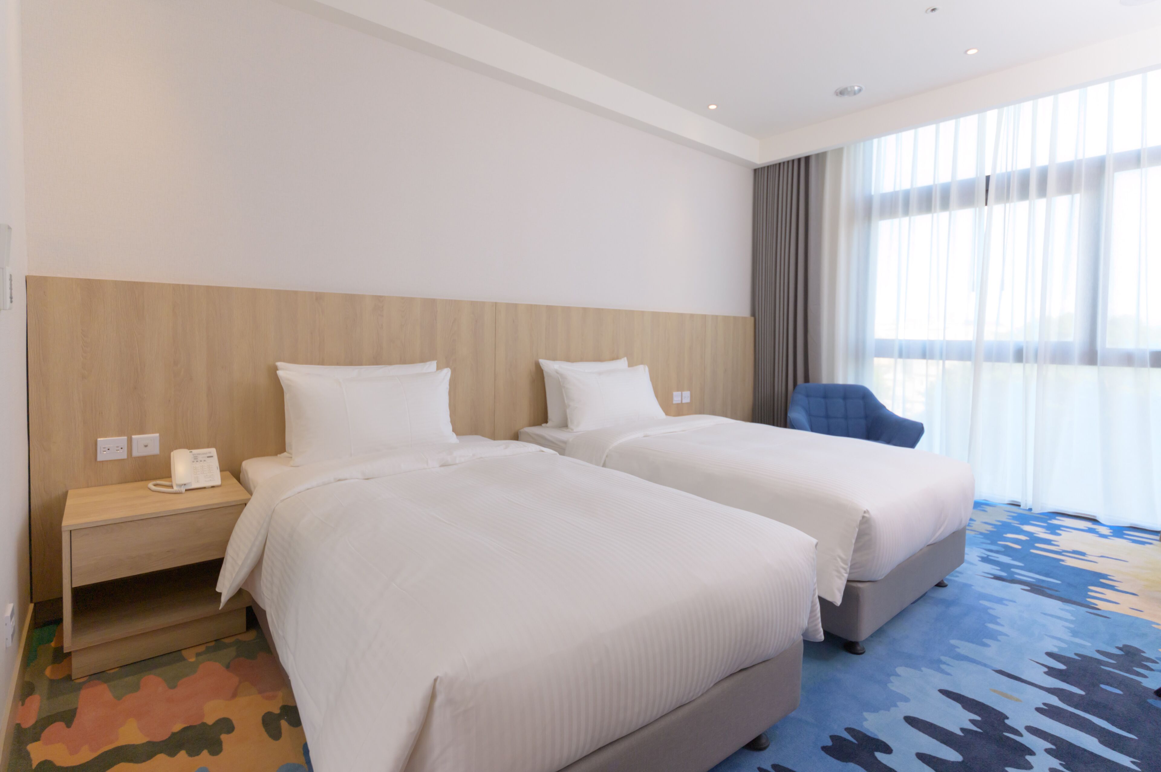 A hotel room with two beds, a nightstand with a phone, a blue armchair, and a large window with curtains.