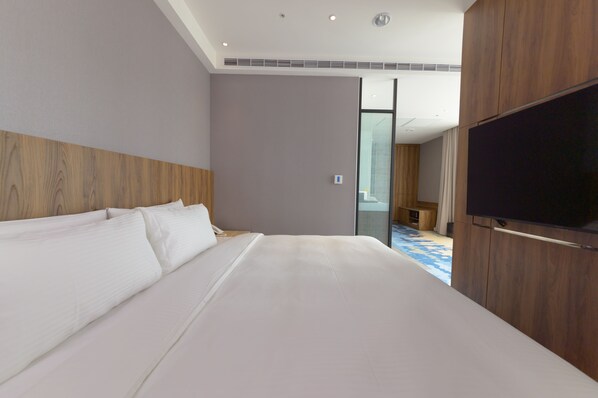 Une chambre d’hôtel moderne dotée d’un grand lit, d’une tête de lit en bois, d’un téléviseur à écran plat et d’une paroi en verre donnant sur la salle de bain.