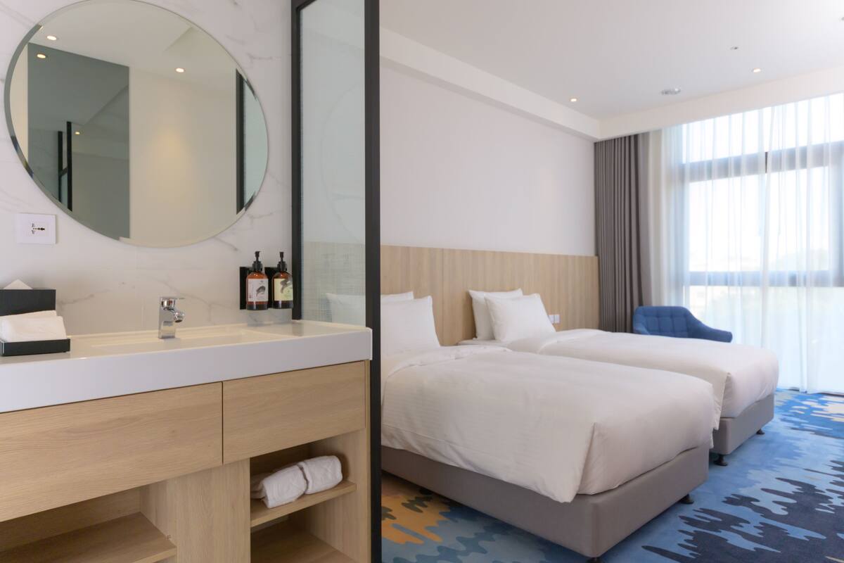 Une chambre d’hôtel avec un miroir rond, un lavabo blanc, une coiffeuse en bois, deux lits, une chaise bleue et une grande fenêtre avec des rideaux.