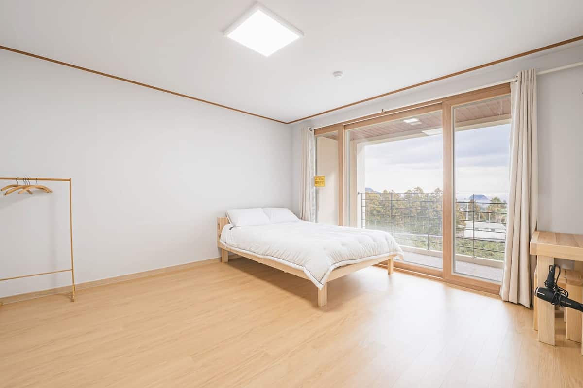 A bedroom with a bed, a wooden floor, a window, and a small table with a bottle.