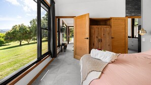 Ein Schlafzimmer mit großen Fenstern, einem Bett, einem Holzschrank und Blick auf eine grüne Landschaft.