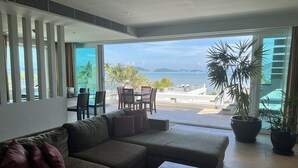 A modern living room with a sofa, a coffee table, and a dining area with chairs and a table. There is a large window with a view of the sea and palm trees. - Stunning Ocean View Studio Apartment (Tambon Rawai)