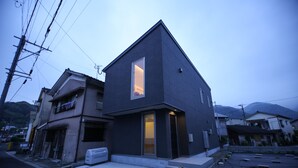 Une maison moderne à deux étages, dotée d’une grande fenêtre au deuxième étage, d’un garage apparent et d’un toit plat.