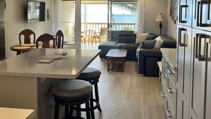 Una cocina moderna con una isla central, taburetes de bar y un comedor. Hay un ventilador de techo, un televisor montado en la pared y vistas al océano a través de puertas corredizas de vidrio.