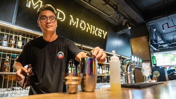 A bartender at a bar with a neon sign reading "MAD MONKEY" in the background. - Mad Monkey Hanoi - Hostel (Hanoi)