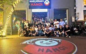 A group of people posing in front of a restaurant named "Mad Monkey Hanoi" with a large sign and a colorful logo on the ground. - Mad Monkey Hanoi - Hostel (Hanoi)