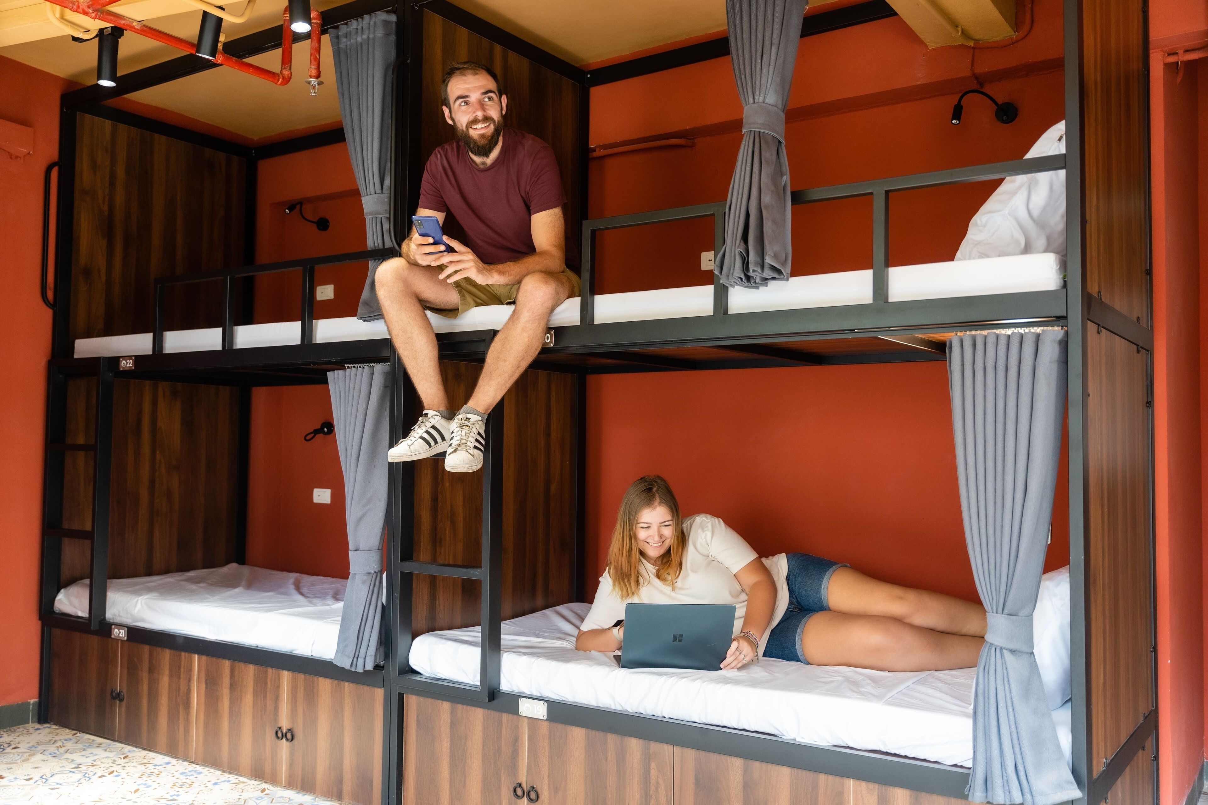 A bunk bed room with a person sitting on the top bunk and another person lying on the bottom bunk using a laptop.