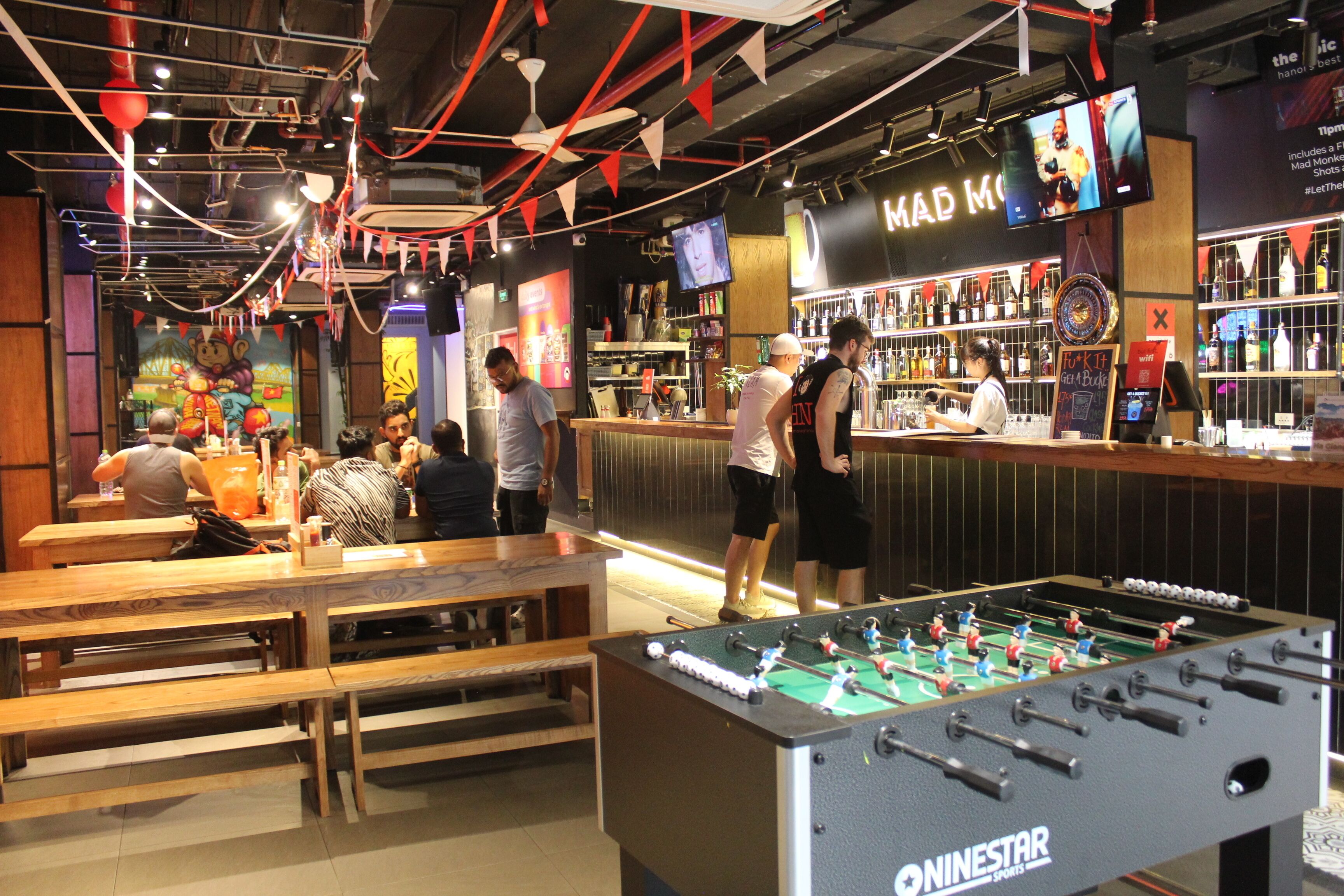 A casual indoor setting with a foosball table, wooden benches, a bar area, and a mural on the wall.