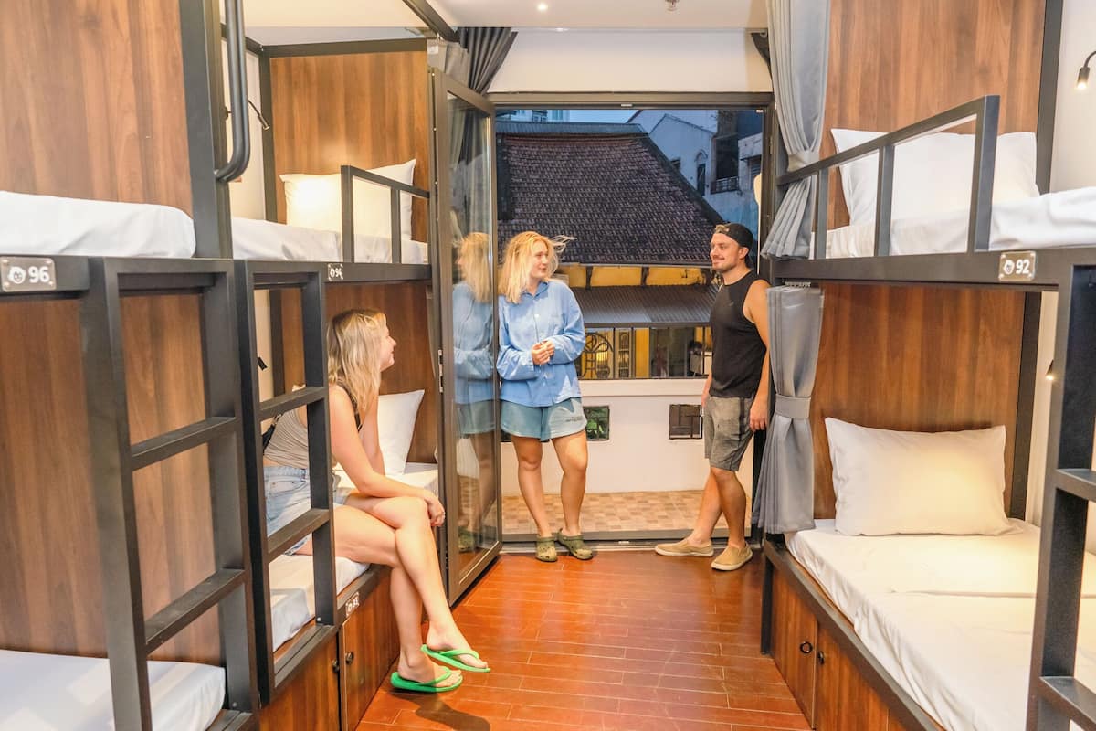 A dormitory room with bunk beds, a wooden interior, and a view of a building outside.