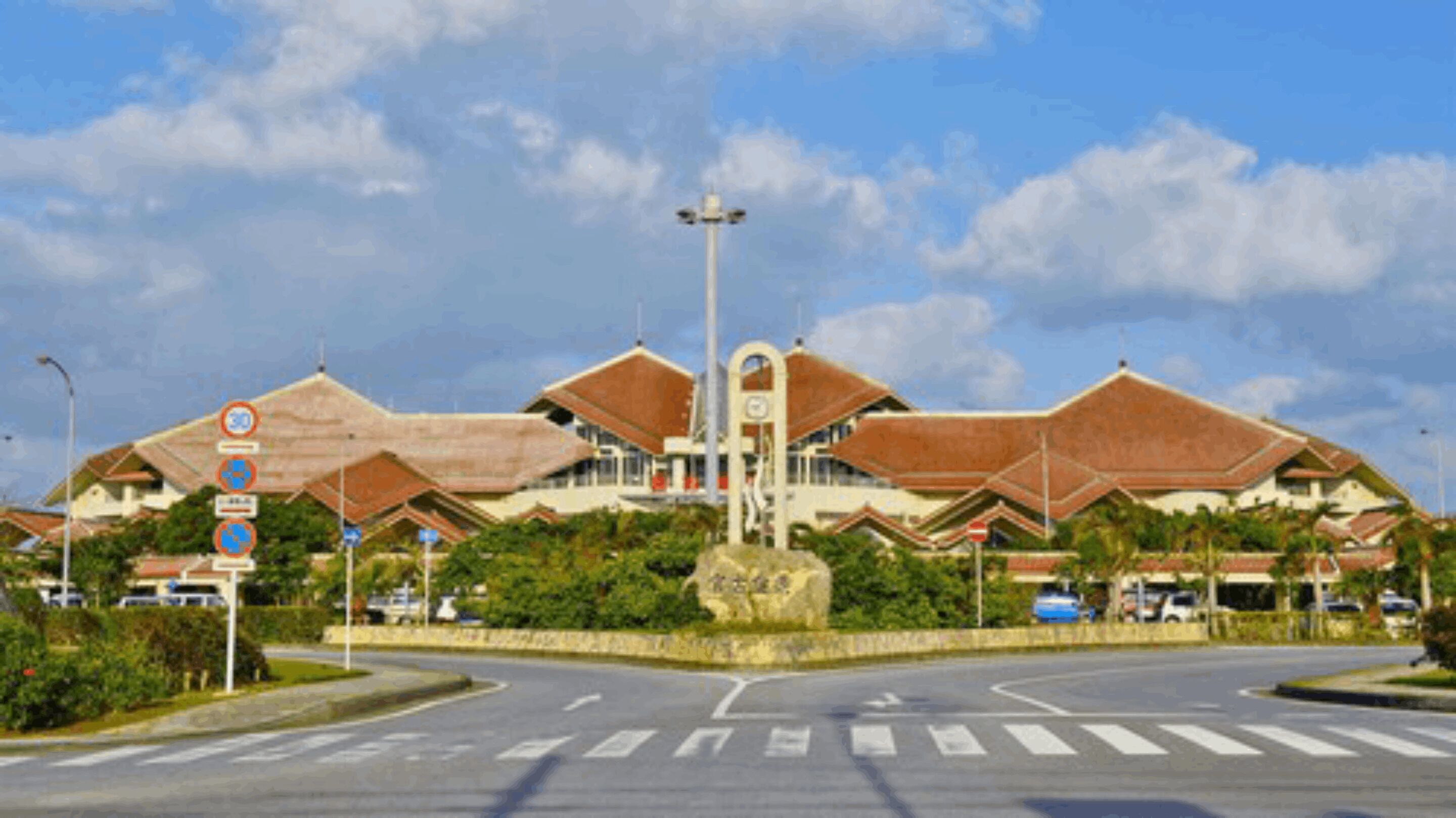 A large building with a central tower and multiple wings, surrounded by greenery and a clear sky.