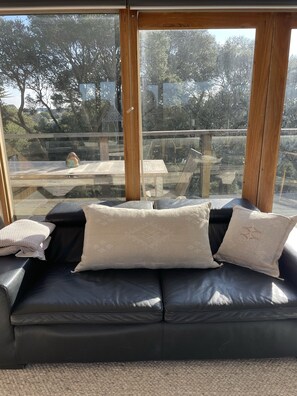 Relax in a spacious room with a large window offering a view of the outdoor area and deck, featuring a comfortable sofa and wooden beams.