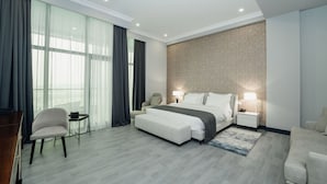 Quarto de hotel moderno com uma cama grande, área de estar com uma poltrona e uma mesa pequena, mesa de cabeceira com abajur e uma janela ampla com cortinas.