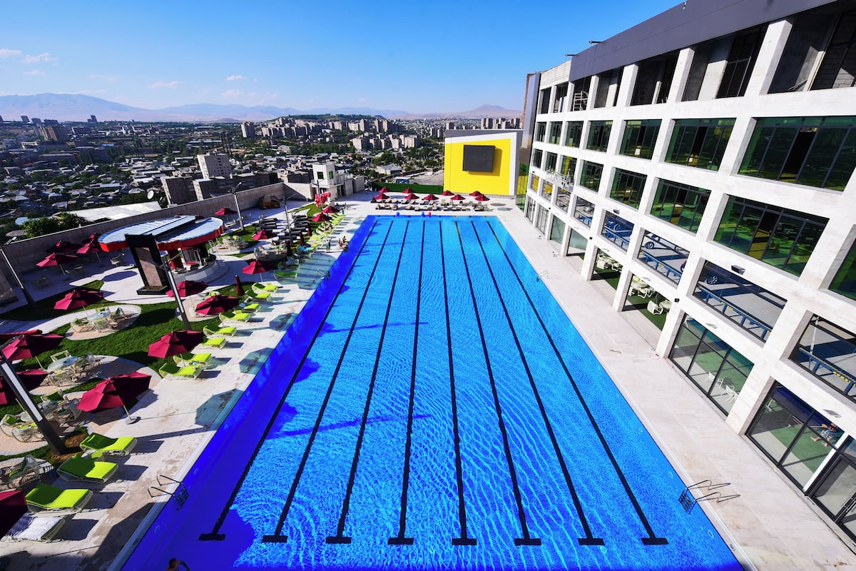A rooftop swimming pool with multiple lanes, surrounded by lounge chairs and umbrellas, overlooking a cityscape.