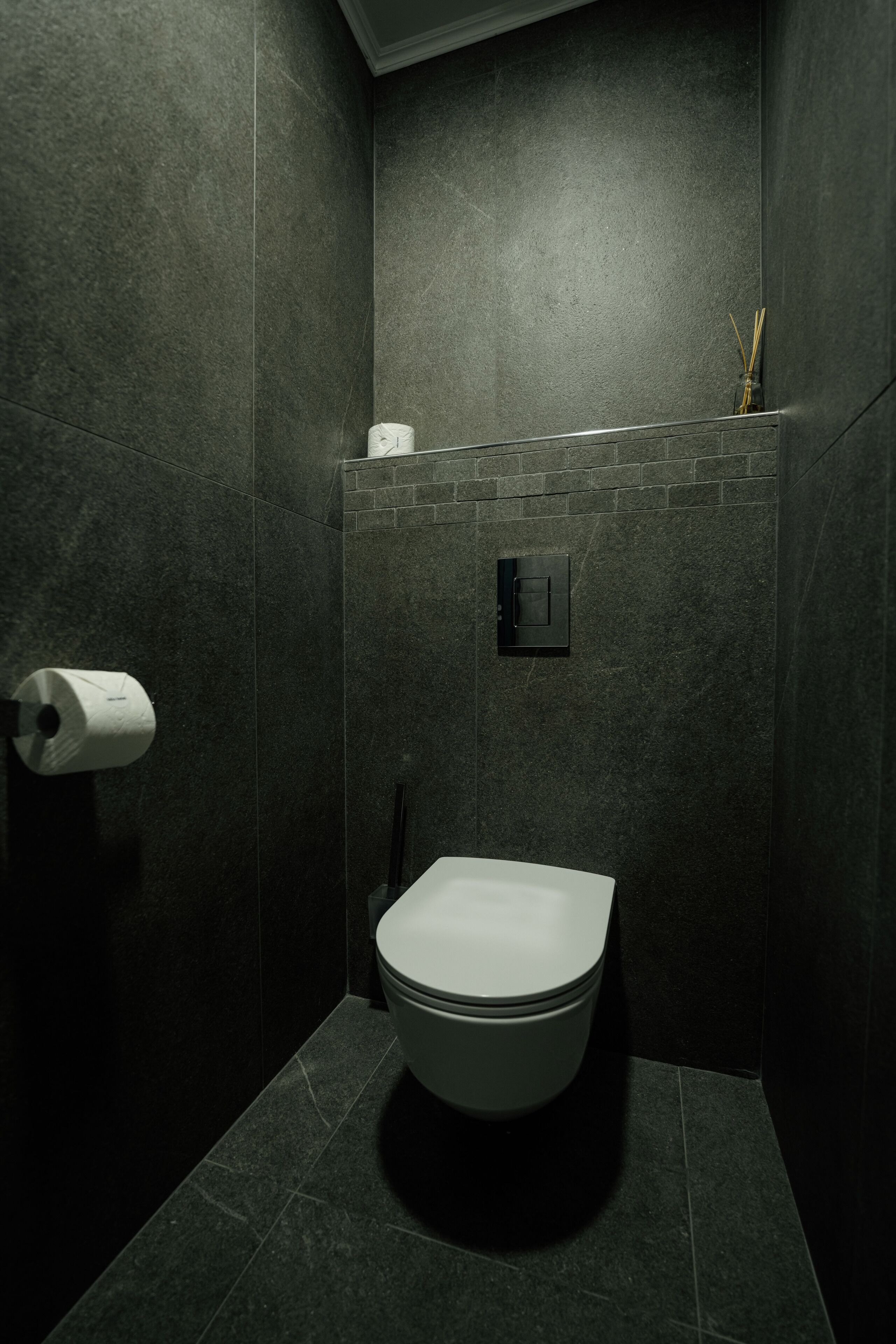 Une petite salle de bain aux couleurs sombres, équipée de toilettes, d’un distributeur d’essuie-tout et d’une petite étagère sur laquelle se trouve une bouteille.