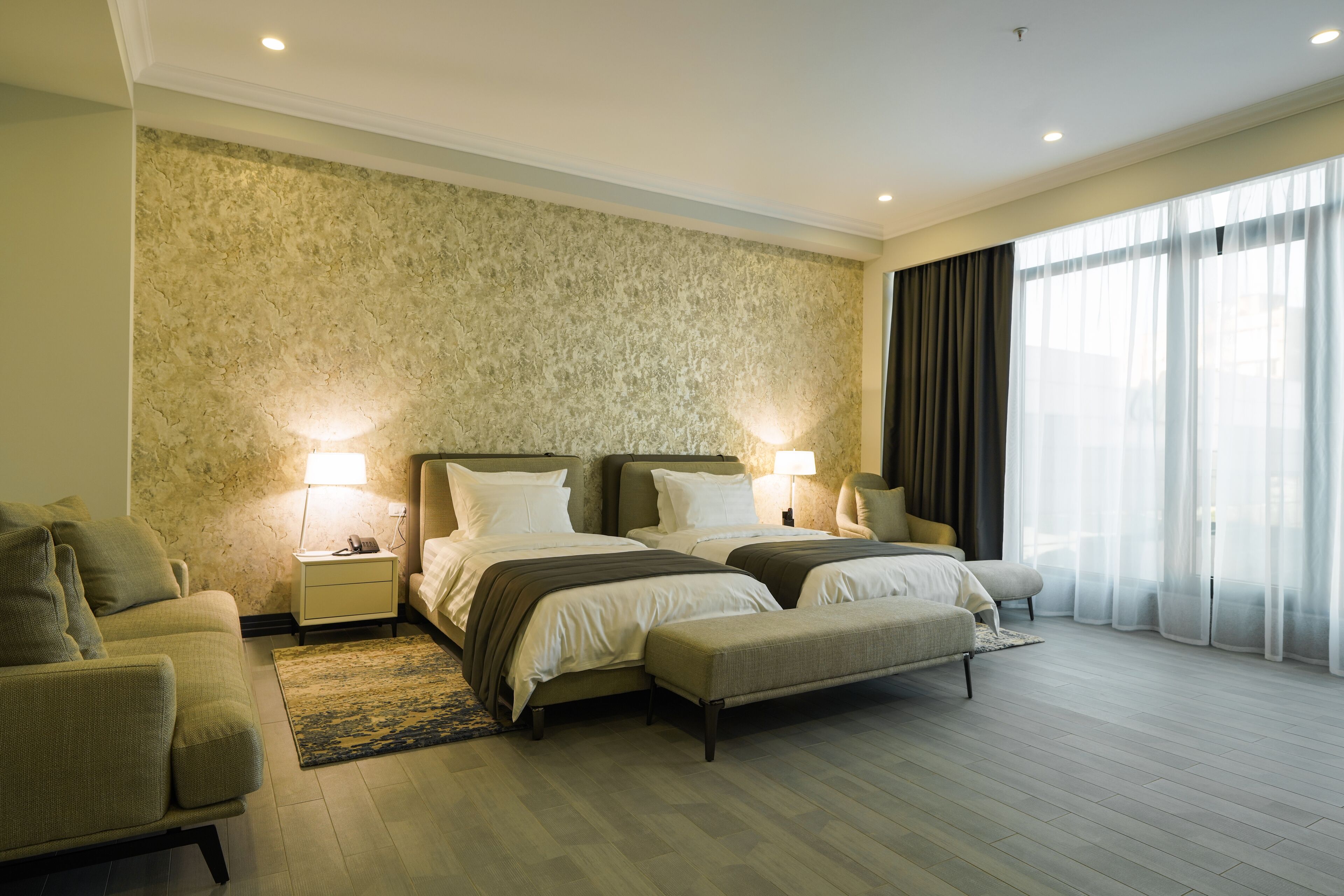 Quarto de hotel com duas camas, um sofá, uma mesa de cabeceira e uma janela grande com cortinas.
