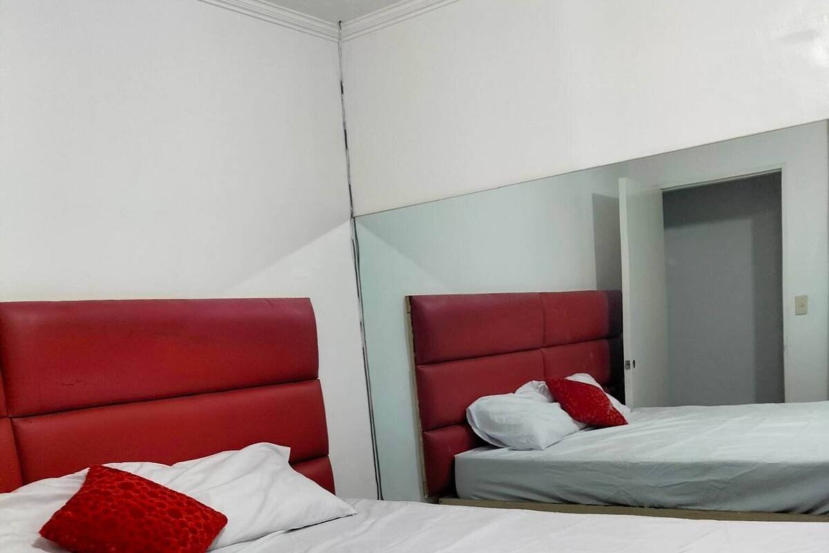 Two beds with red headboards and white bedding in a room with white walls and a mirror reflecting part of the room.