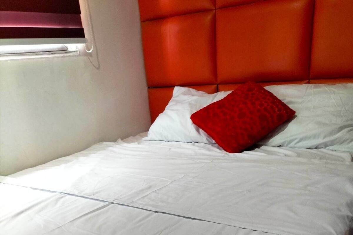 A bed with white bedding and a red pillow, a window with blinds, and an orange headboard.