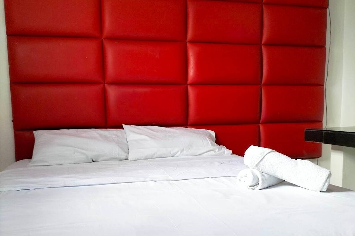 A bed with white linens and pillows, a red padded headboard, and a black nightstand with a white towel.
