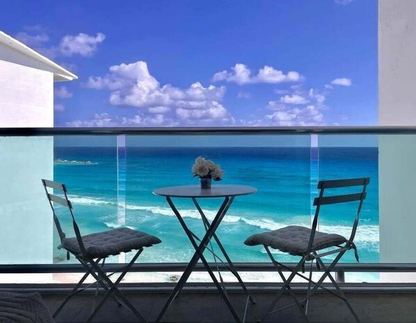 Un balcon avec deux chaises et une petite table, d’où l’on peut admirer une mer bleue et limpide et un paysage lointain.