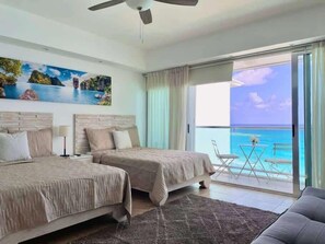 Une chambre d’hôtel avec deux lits, un ventilateur de plafond, un tableau représentant un paysage tropical, un balcon avec vue sur l’océan et une petite table avec des chaises.