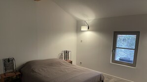 Ein Schlafzimmer mit Bett, Nachttisch, Lampe und Fenster mit Blick auf eine Steinmauer.