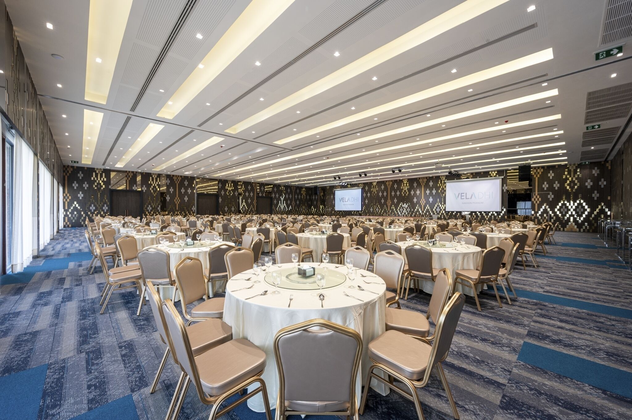 A conference room with round tables, chairs, and a projector screen displaying "VELADHI".