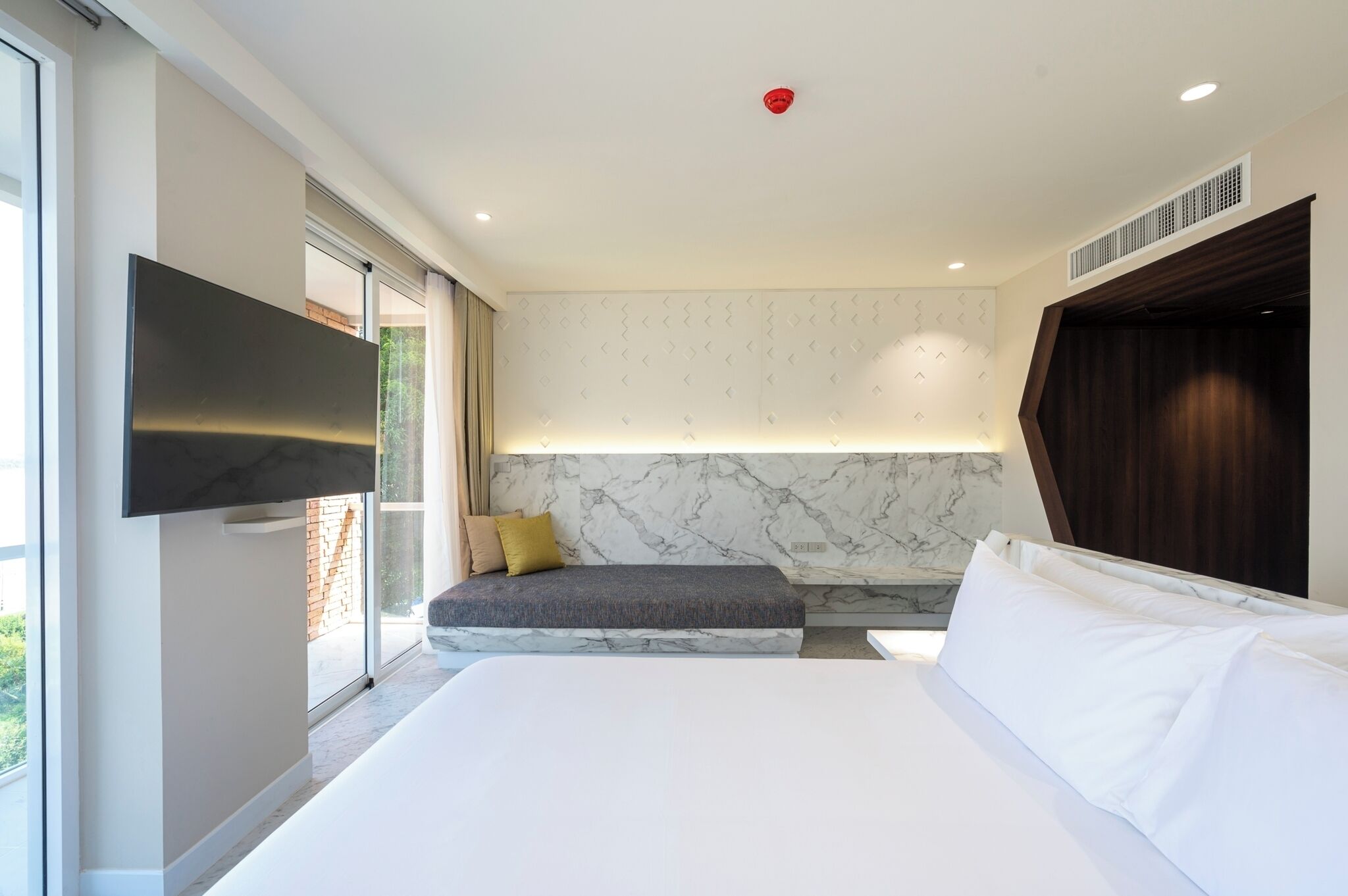 A modern bedroom with a large bed, a flat-screen TV mounted on the wall, a seating area with cushions, and a marble-patterned wall.