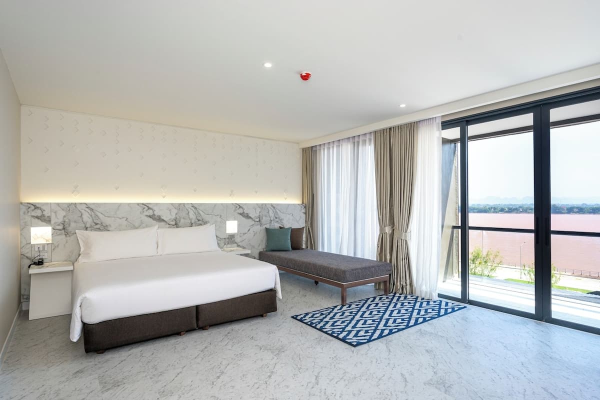 Habitación de hotel moderna con una cama grande, un banco, una pared de mármol y vistas a un río.