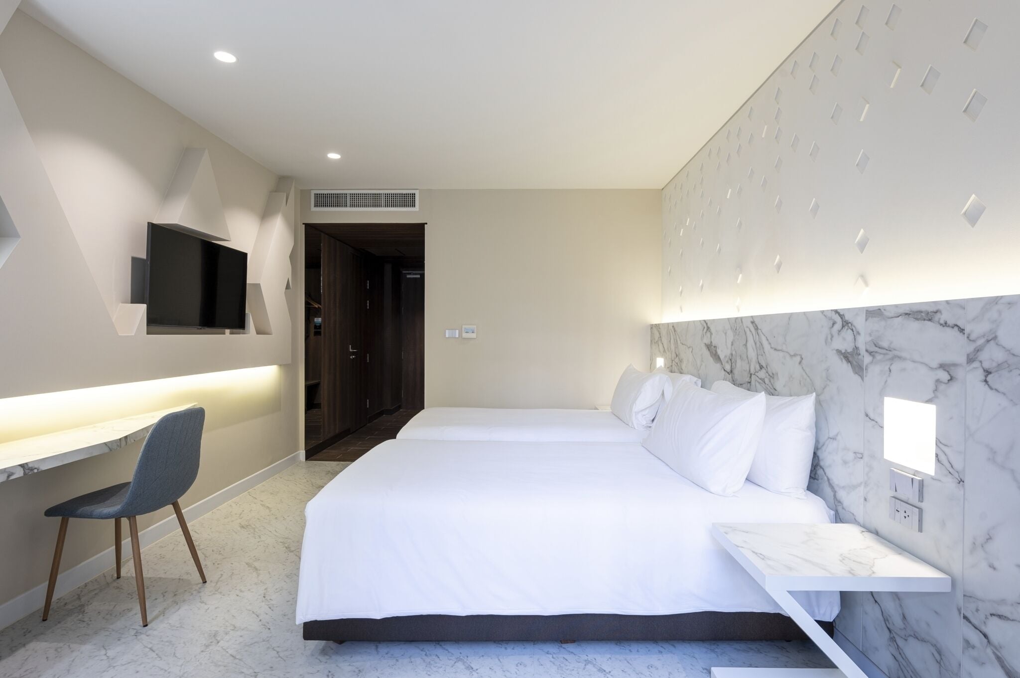 A modern hotel room with a large bed, a desk with a chair, a flat-screen TV mounted on the wall, and a marble-patterned headboard.