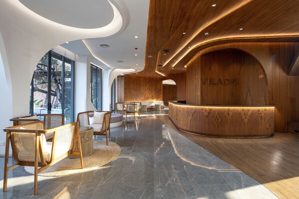 Un área de recepción moderna con un mostrador de madera, zona de estar con sillas de mimbre y ventanas amplias que ofrecen vistas al exterior.