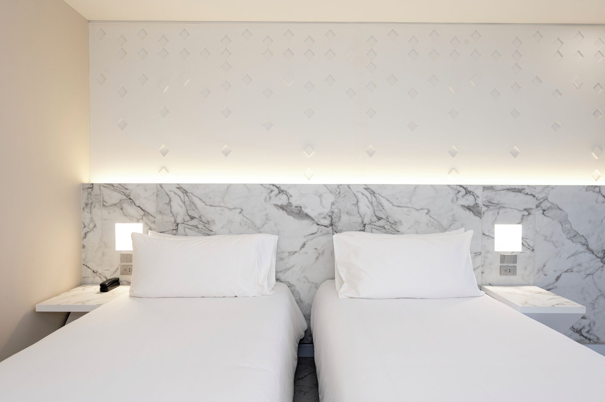 Een hotelkamer met twee bedden, een marmeren hoofdbord, bedlampjes en een behang met een patroon.