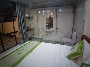 Une chambre d’hôtel dotée d’une douche en verre, d’un lit avec une literie verte et blanche, et offrant une vue sur l’extérieur à travers la porte en verre.