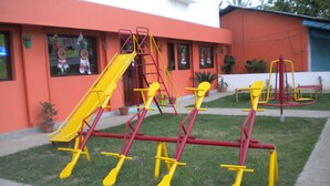 A playground with yellow and red equipment, including swings and a slide, in front of a building with windows and potted plants. - Rains Inn Eco Hotel (Diphu)