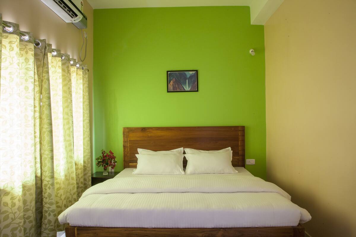 Cama com roupa de cama branca, cabeceira de madeira e parede verde com um quadro.