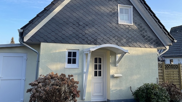 Une maison de plain-pied avec un toit recouvert de tuiles grises, une porte blanche et deux fenêtres.