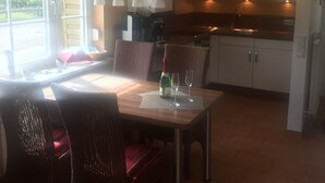 Une salle à manger avec une table dressée pour deux personnes, des chaises, une bouteille de vin et des verres. Il y a une fenêtre avec des rideaux, une horloge au mur et une cuisine en arrière-plan.