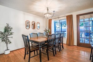 A dining room with a wooden table and chairs, a chandelier, a potted plant, and a view through a window with curtains.