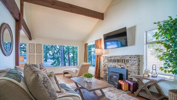 A cozy living room with a fireplace, comfortable seating, and large windows offering a view of trees. - Fairyland Cottage: Waterfront+Hot Tub+Beach+Clam (Poulsbo)