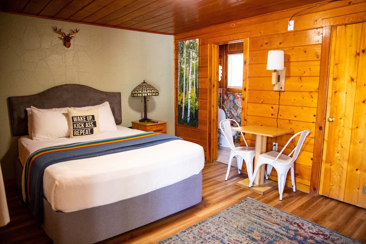 A wooden cabin room with a bed, a table with chairs, a lamp, and a wall-mounted light fixture.