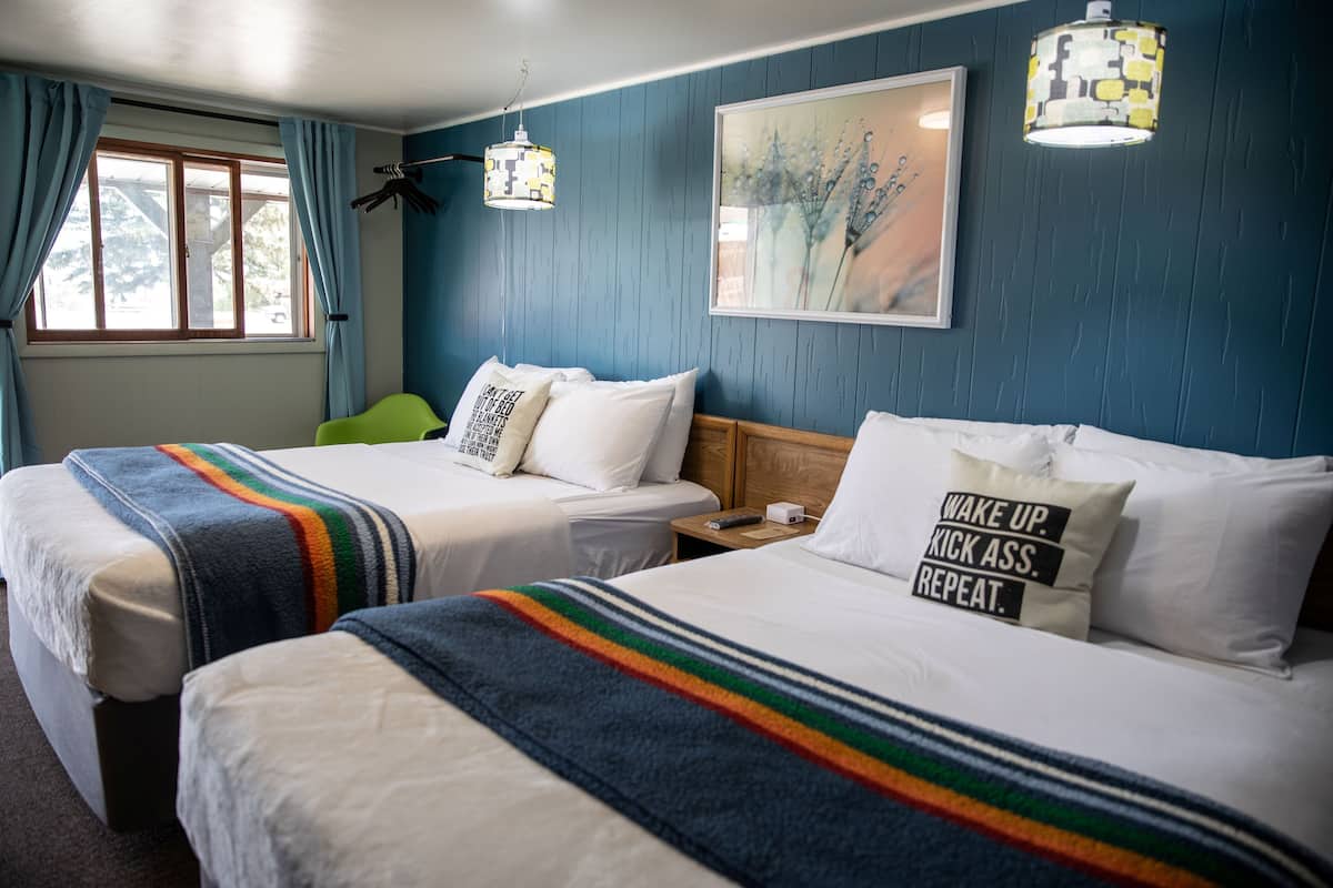 A hotel room with two beds, a window with curtains, a painting on the wall, and a lamp.