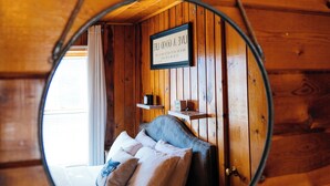 Een ronde spiegel weerspiegelt het interieur van een houten hutje, met een bed, kussens en een bord aan de muur.