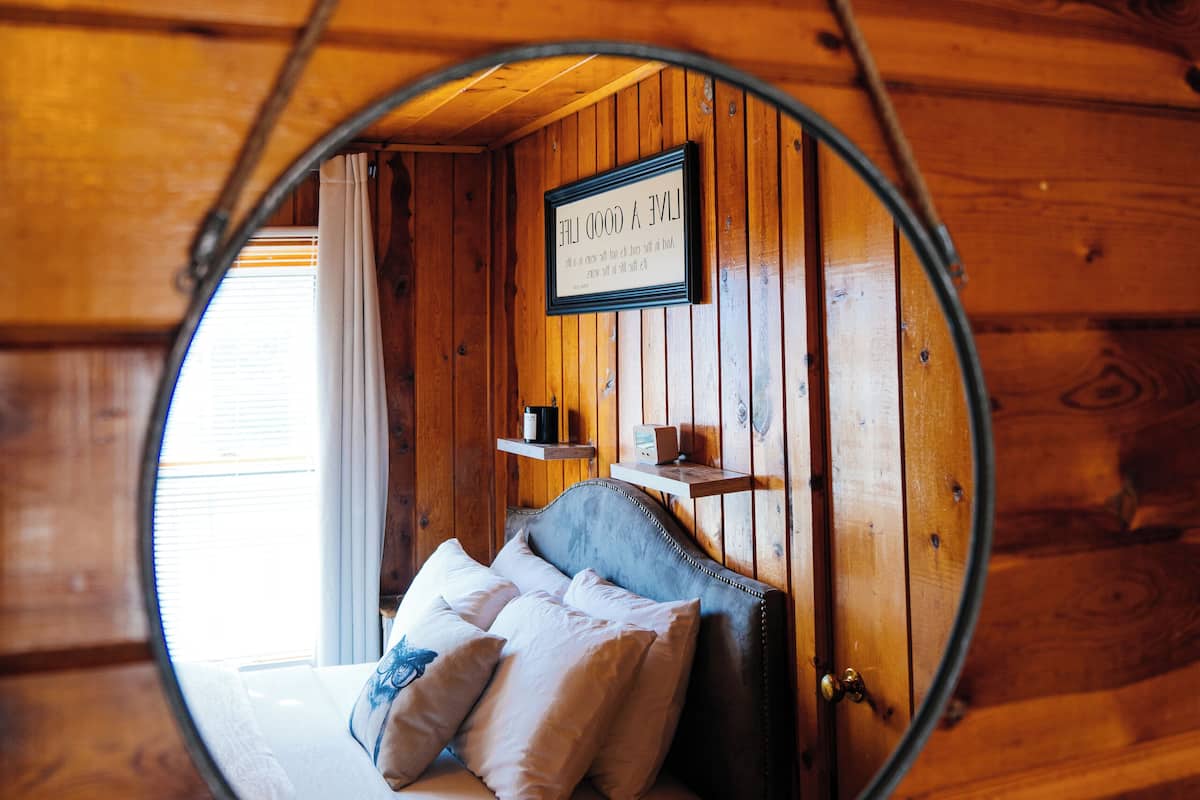 A circular mirror reflecting a wooden cabin interior with a bed, pillows, and a sign on the wall.