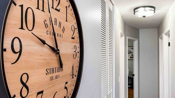 A large wall clock with a wooden face and black numbers, mounted on a white wall in a hallway with a patterned rug and a door leading to another room. - Mindfully Mellow | 3Bd 1Bath - Berwick (Lansing)