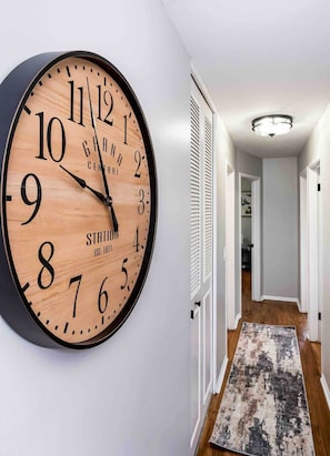 Un reloj de pared grande con esfera de madera y números negros, colgado en una pared blanca en un pasillo con un tapete estampado y una puerta que da a otra habitación.